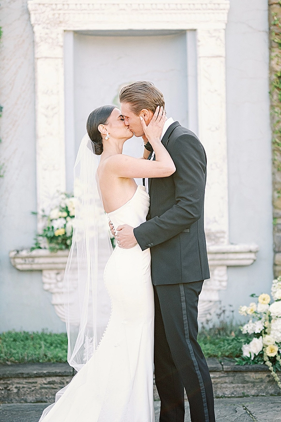 Wedding kiss portrait of bride and groom kissing, her veil flowing as she holds his face by a stucco wall niche with greenery