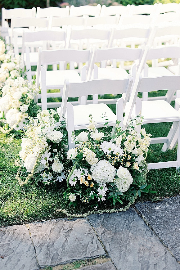 Ceremony aisle decor with outdoor ceremony aisle flowers lining a stone walkway, framed by white folding chairs on a garden lawn.