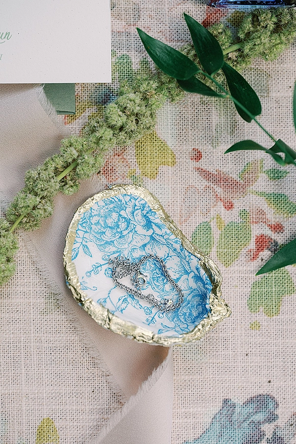 Wedding jewelry flatlay with silver bracelet on blue floral trinket dish beside invitation, linen napkin, and greenery on patterned fabric tablecloth