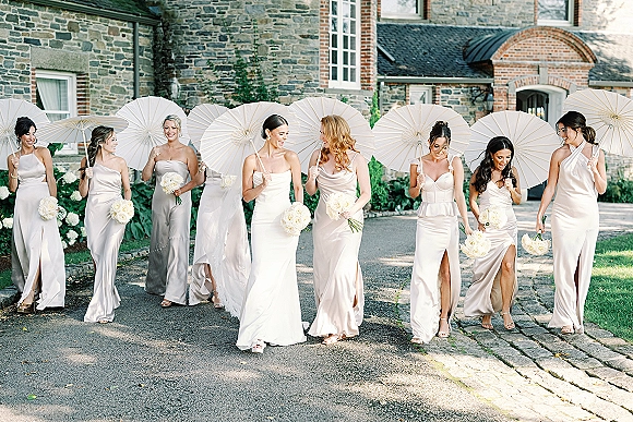 Bridesmaids portrait with white parasols, carrying white rose bouquets in satin dresses on a manor driveway before a stone facade