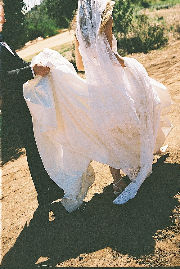 Wedding dress detail with lace wedding veil flowing as groom in black tuxedo lifts the train on a sunlit dirt path with greenery