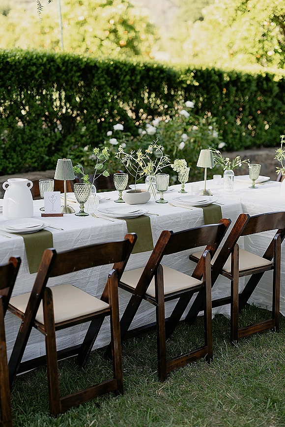 Reception tablescape with olive green runner, green goblets, white plates, bud vases and small lamps on a long table in sunny garden lawn
