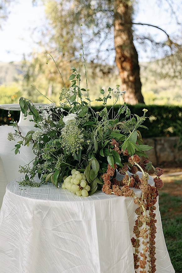 Wedding floral arrangement green and white centerpiece with airy greenery and grapes on a white tablecloth in a sunlit garden setting