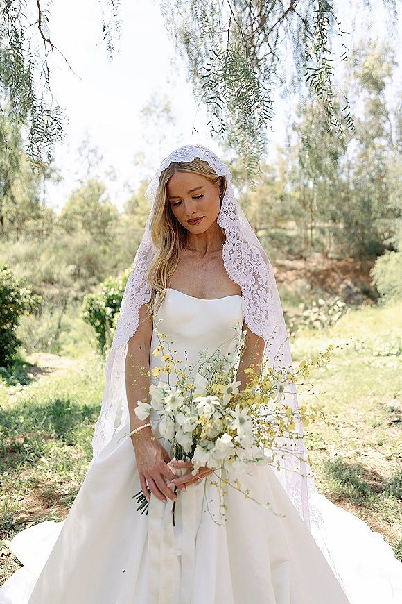 Bridal portrait of a bride in a mantilla veil holding a white and yellow wildflower bouquet in a sunlit garden setting