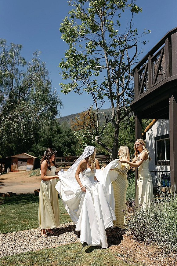 Bride getting ready as bridesmaids help fasten her wedding dress on a wooden deck, veil trailing with mountains and blue sky behind