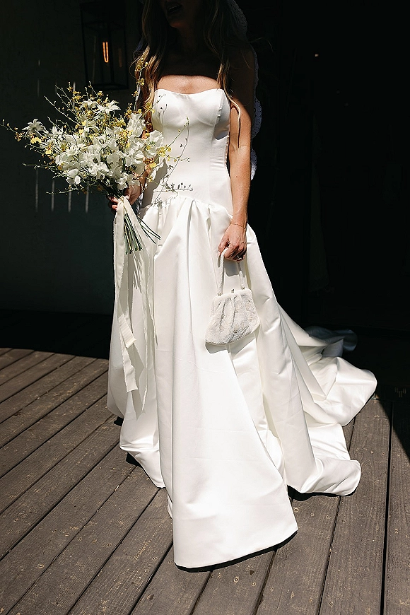 Bridal portrait of a bride in a strapless satin wedding dress holding a white bouquet with ribbon, long veil, and fuzzy purse by a dark wall