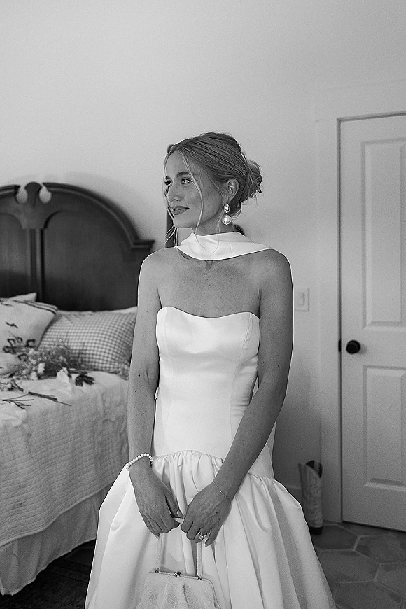 Bridal portrait of a bride in a strapless wedding dress with neck scarf and pearl drop earrings, holding a handbag in a bedroom