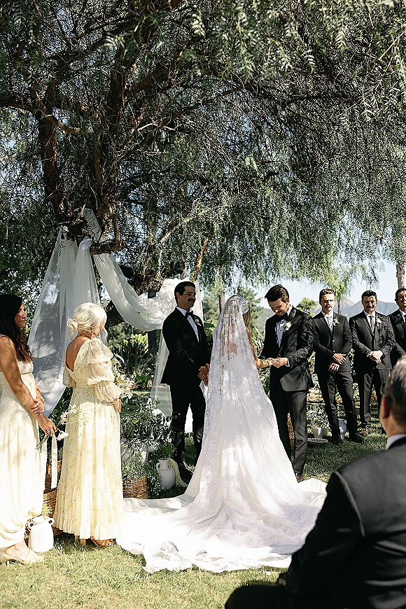 Wedding ceremony as couple exchanges rings beneath a large tree, bride’s lace veil and long train flowing beside tuxedoed groomsmen