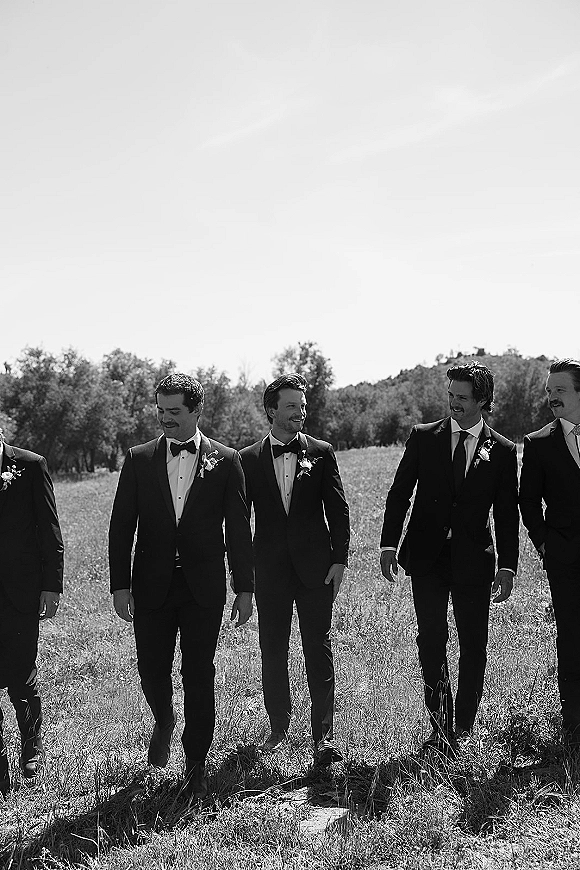 Groomsmen portrait of men in tuxedos with boutonnieres walking together across a grassy field with trees and hills behind them