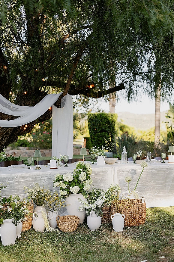 Reception tablescape with white linen head table, green goblets, ceramic vases of white florals, candles and string lights under a large tree canopy