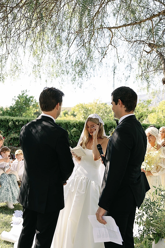Wedding vows as bride reading vows beside groom in tuxedo, holding vow book with veil flowing at a sunlit garden ceremony
