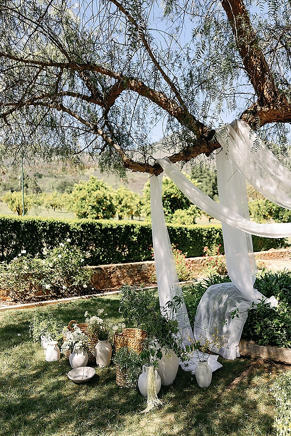 Ceremony backdrop with white draping fabric and hanging textiles on tree branches, framed by green-and-white florals in vases and baskets on lawn