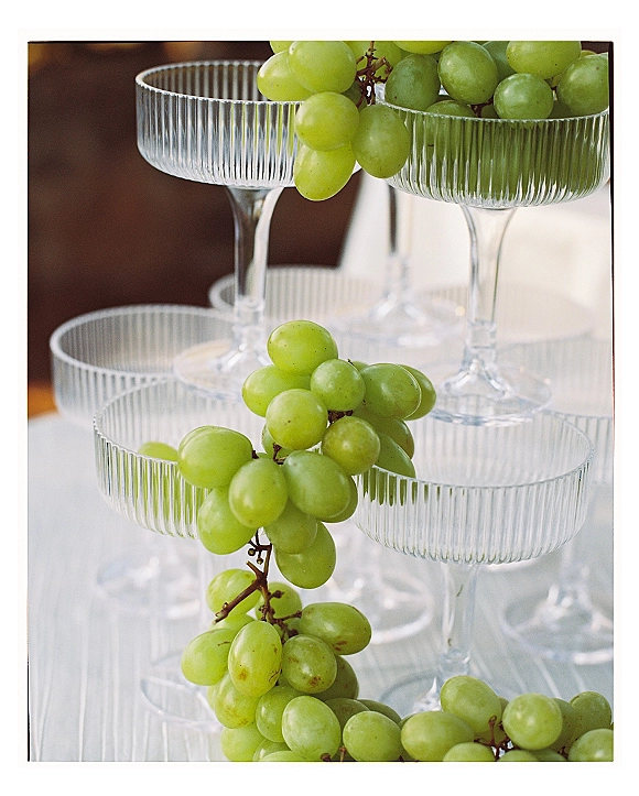 Champagne tower of coupe glasses with green grapes nestled between tiers on a white tablecloth, set against a softly blurred interior