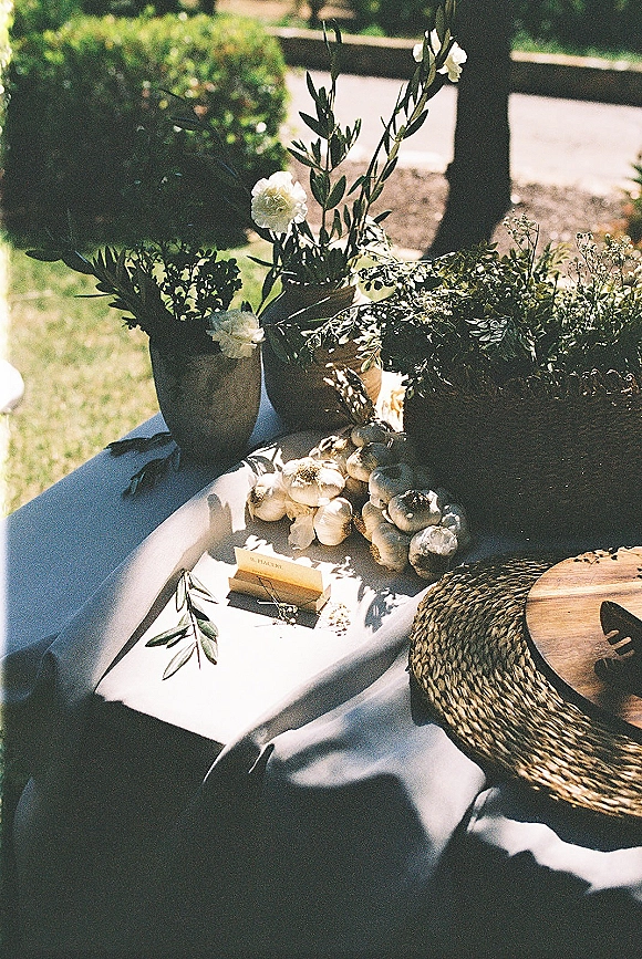 Reception tablescape with outdoor wedding table decor, white flowers and greenery in clay vases, garlic bulbs on linen, set on a lawn by a wooden fence