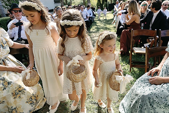 Flower girls in flower girl dresses walk down an outdoor ceremony aisle, holding wicker baskets with white ribbon bows among guests and chairs