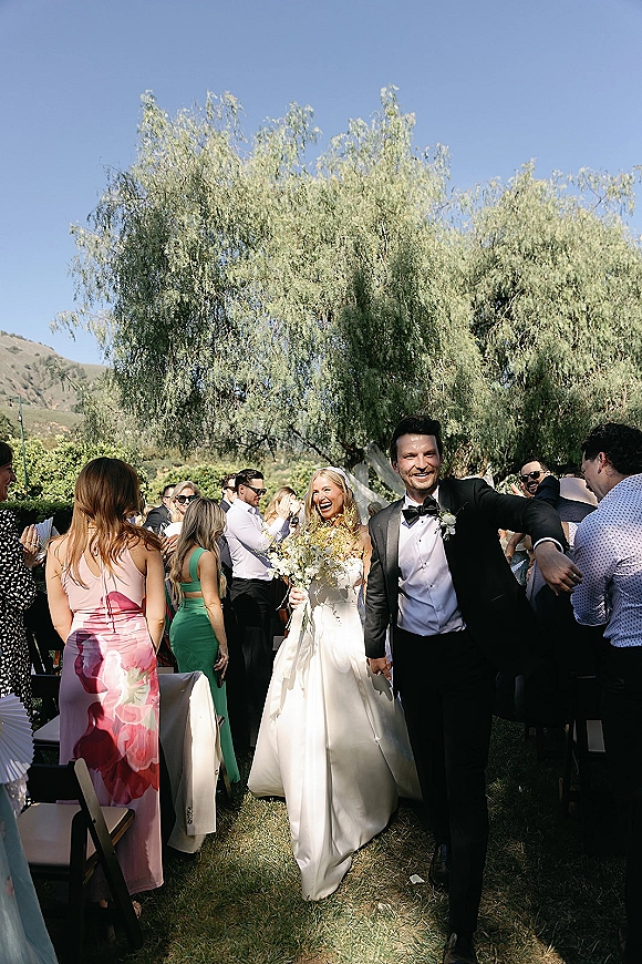 Wedding recessional as bride and groom walk the aisle, bride holding bouquet, veil flowing as guests cheer on a sunny lawn with hills