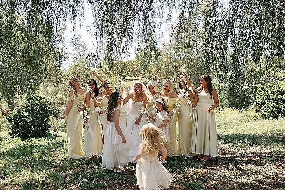 Bridesmaid group photo with bridesmaids in neutral dresses surrounding the bride, holding bouquets with ribbons in a sunlit garden setting