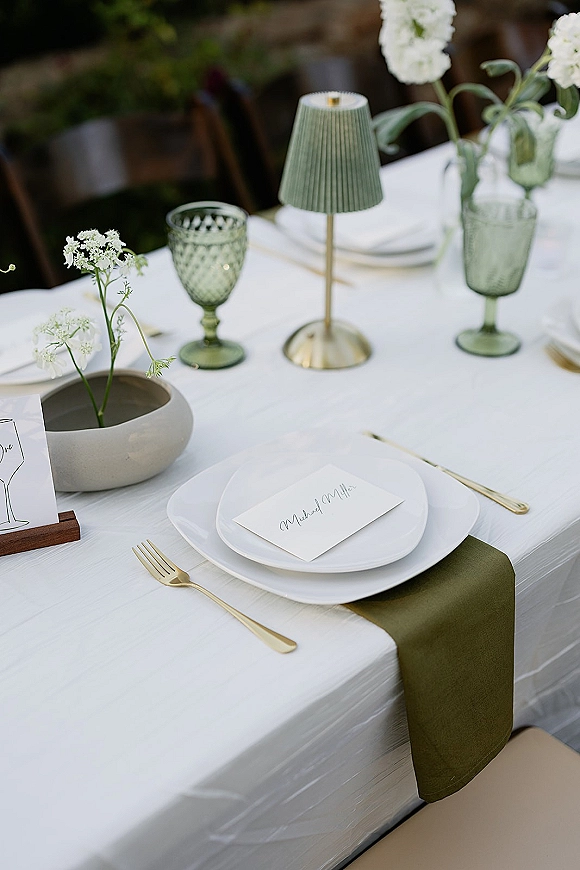 Reception tablescape with wedding place setting, green runner, gold flatware, green goblets, and a small lamp beside white flowers
