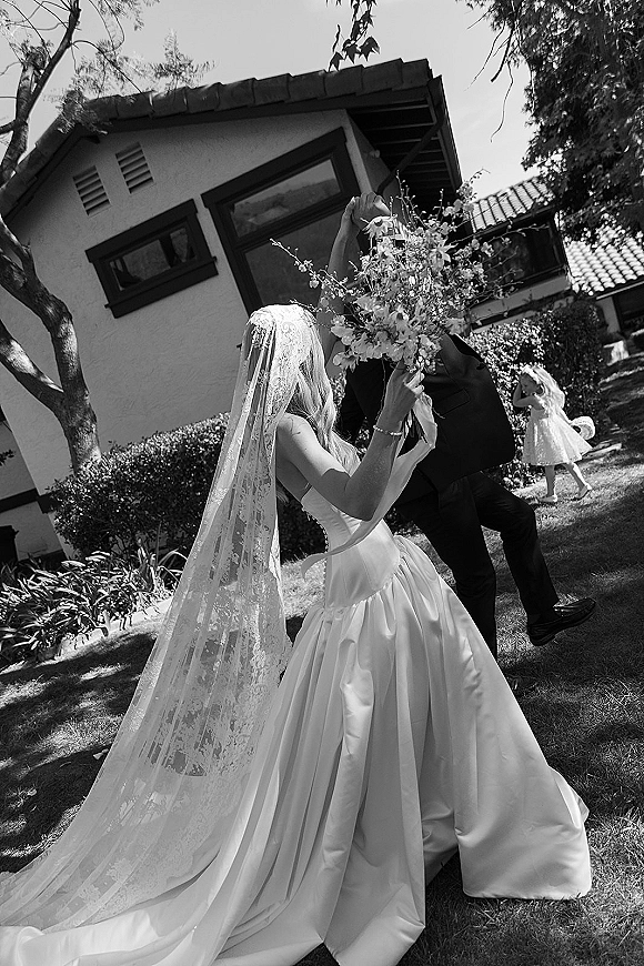 Wedding kiss portrait of bride and groom kiss as he lifts her, lace veil flowing and bouquet in hand on a green garden lawn