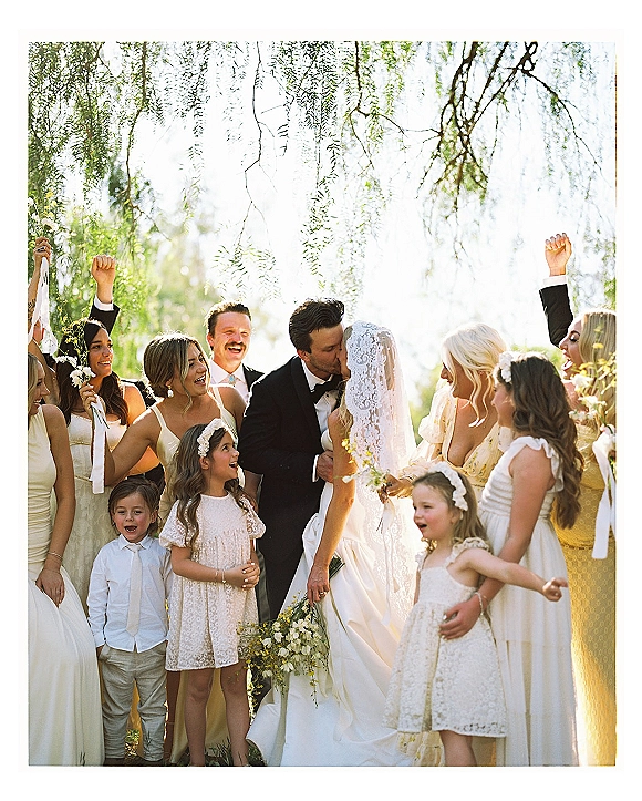 Wedding kiss moment as the newlyweds kiss in sunlight, bridal party and flower girls cheering with ribbons in an outdoor garden setting