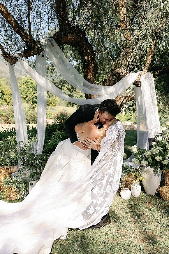 Wedding kiss as groom dips the bride, her lace veil flowing beside draped fabric and white florals on a sunlit garden lawn