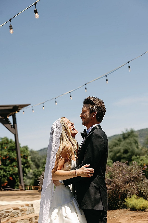 Couple portrait of bride and groom embrace, bride laughing in veil with string lights on a wooden pergola against blue sky and hills
