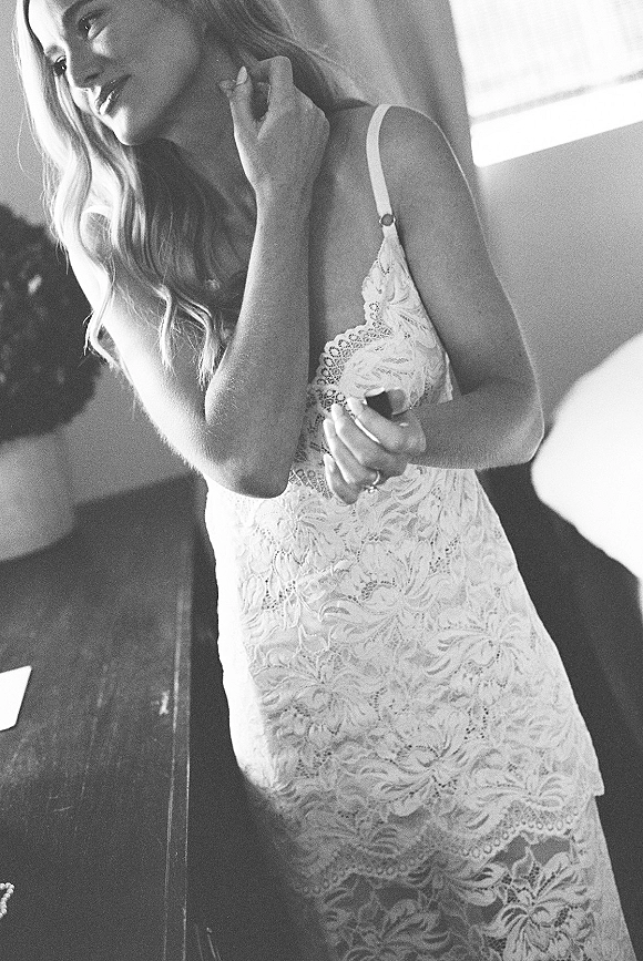 Bridal portrait of a bride in a lace wedding dress adjusting her hair, showing an engagement ring in soft window light by a dresser
