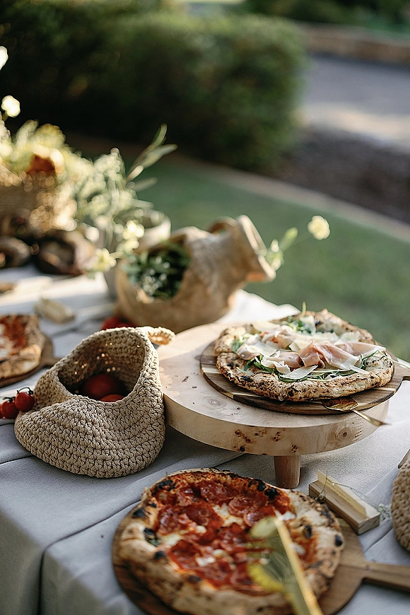 Wedding catering spread with pizza wedding catering on wood boards, paper bags, and greens on a low table on an outdoor lawn