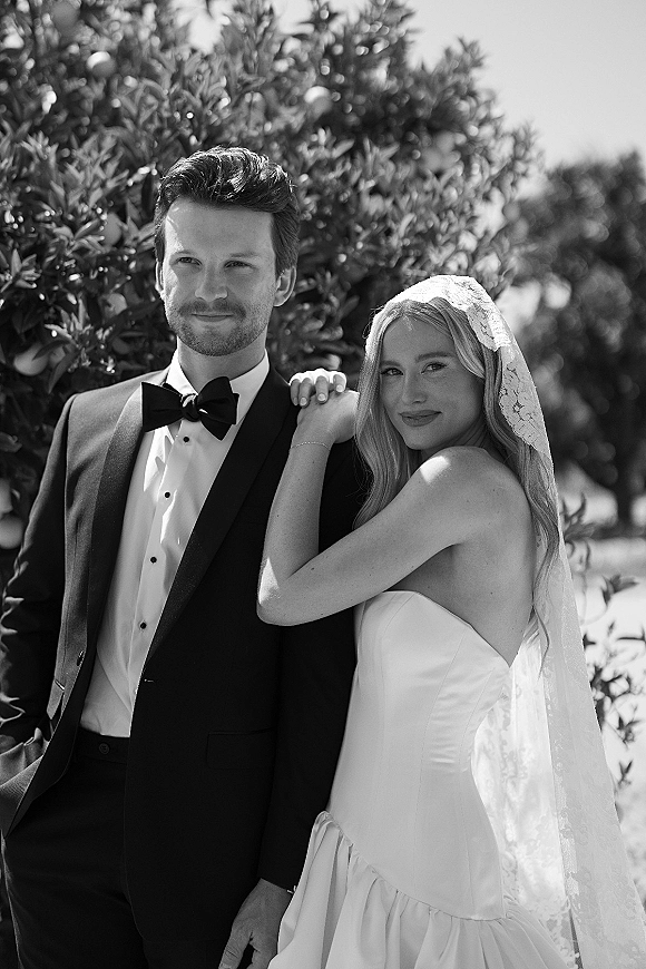 Couple portrait in a black and white wedding portrait style, bride in strapless dress and lace veil leaning on groom in tuxedo beneath sunlit trees