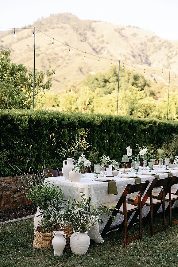 Reception tablescape with an outdoor reception table, olive green runner, green goblets, white florals, and string lights with mountains beyond