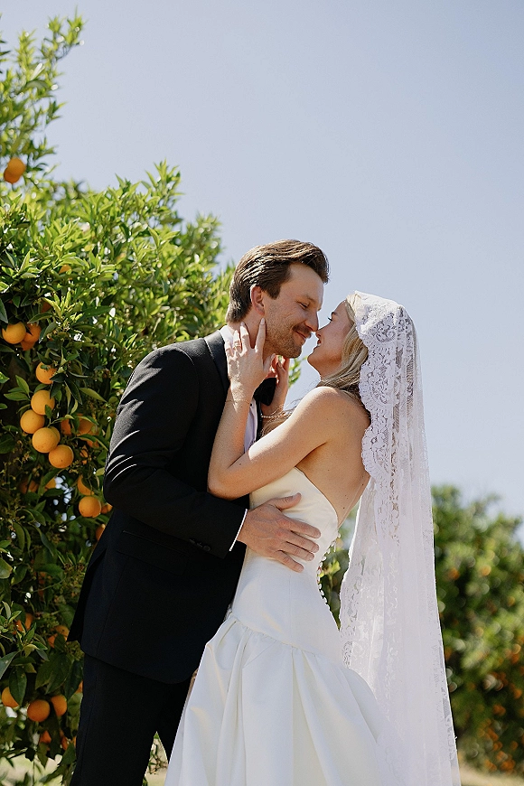 Wedding kiss portrait of bride and groom kissing, her lace veil flowing as they embrace in an orange grove under blue sky