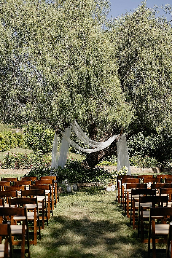 Outdoor ceremony setup with tree wedding altar, white draped fabric, wood chairs, aisle runner, and lanterns beneath a large tree on lawn