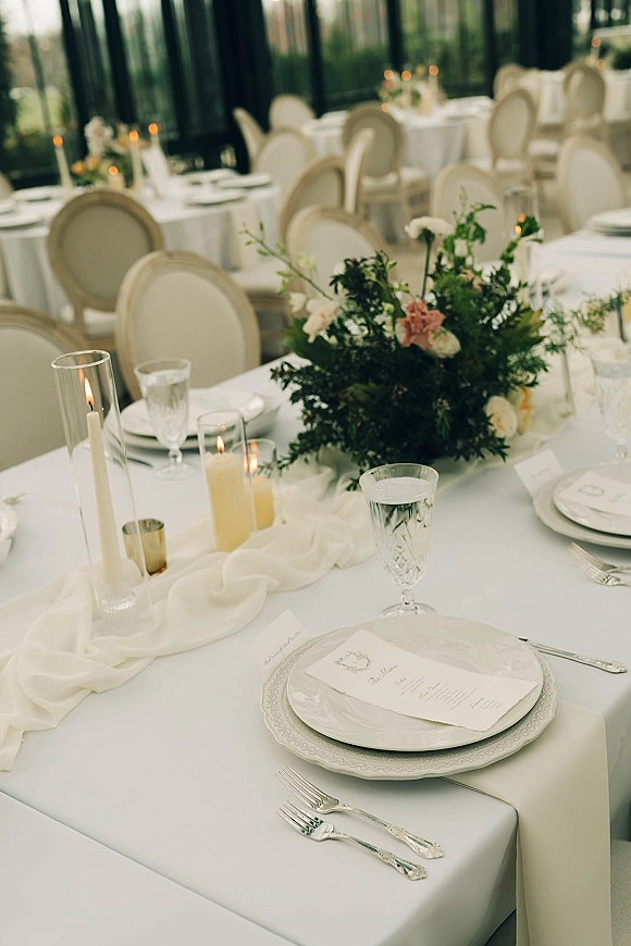 Reception tablescape with a wedding table setting, white linens, greenery and floral centerpiece, taper candles, and glassware by tall windows