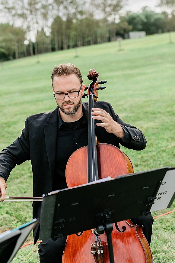 Wedding musician in a black suit plays cello with bow and sheet music on a stand in a grassy field with trees behind