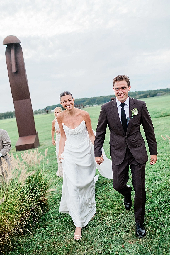 Recessional moment as bride and groom walking hand in hand, bride laughing in strapless gown and veil, black tie suit in grassy field under cloudy sky