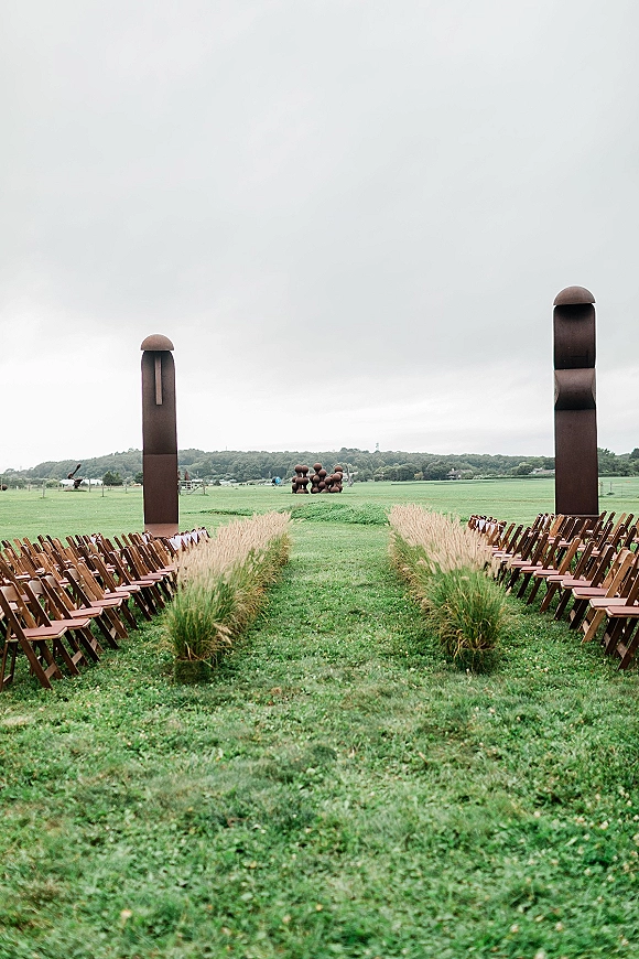 Outdoor ceremony setup with wood folding chairs flanking a grassy aisle, leading to modern altar columns in an open field under cloudy sky