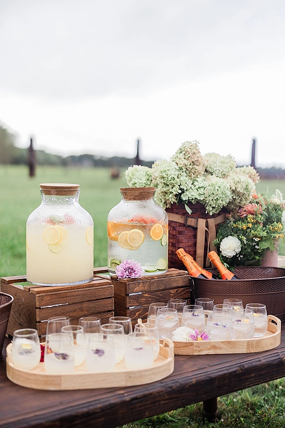 Wedding drink station with glass dispensers and citrus slices, wooden crates, tumblers, and hydrangea accents set in a field by fence posts