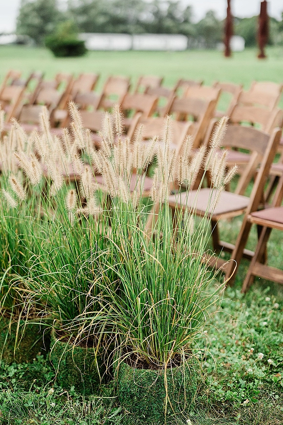 Ceremony aisle decor with outdoor ceremony chairs, greenery and ornamental grass markers along a lawn aisle by trees and a white fence