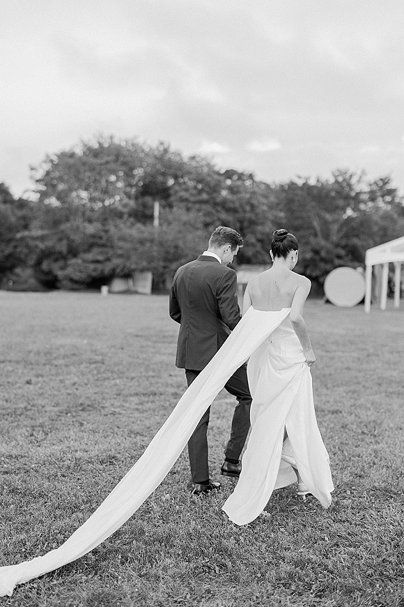 Couple portrait of bride and groom walking away on a lawn, bride holding her dress train and veil trailing near a tent under trees