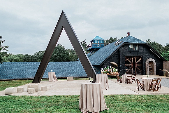 Cocktail hour setup with wedding cocktail tables in taupe linens, candles and greenery, plus escort card display on a concrete patio by a barn and metal sculpture