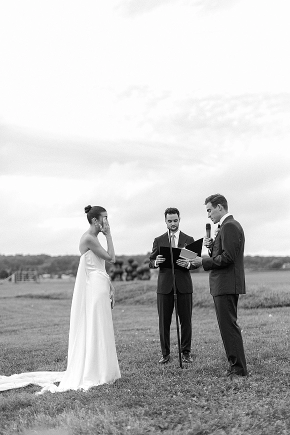 Wedding vows as groom reads paper vows into a handheld microphone beside bride in strapless dress, in an open field under cloudy sky