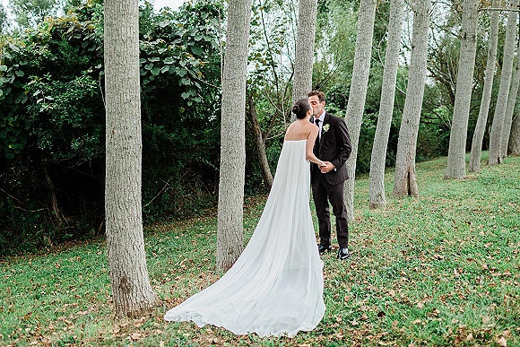 Wedding kiss portrait of bride and groom kissing, holding hands on a grassy tree grove lawn, her strapless dress with long train trailing behind