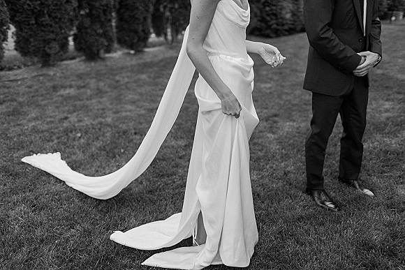 Wedding couple portrait in black and white, bride holding her dress train beside groom in black suit on a garden lawn with trees