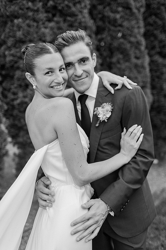 Couple portrait in black and white wedding portrait style, bride in strapless dress and pearl earrings embracing groom in suit amid garden greenery