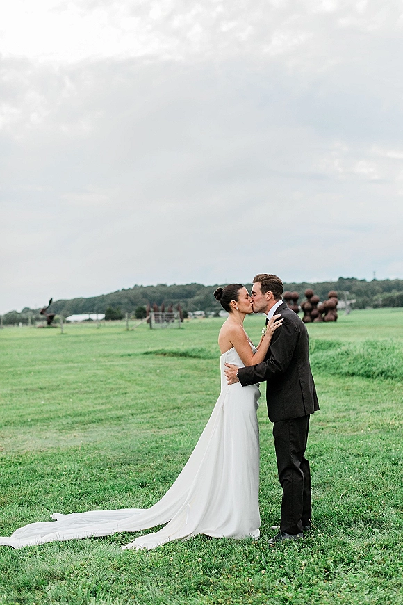 Wedding kiss portrait of bride and groom kiss in a grassy field, her strapless wedding dress with train against a cloudy sky