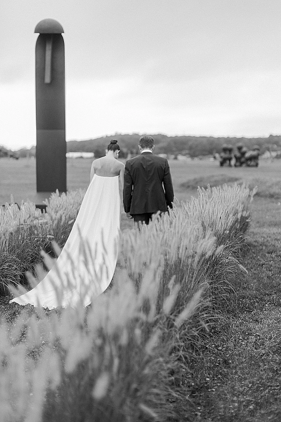 Couple portrait in a black and white wedding photo, newlyweds holding hands from behind in a grassy field with rolling hills and sculpture