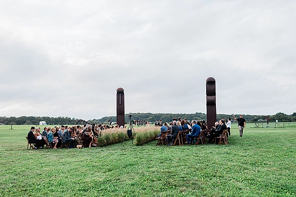Outdoor wedding ceremony with ceremony seating layout of wood folding chairs facing a wide grass aisle in an open field under cloudy skies