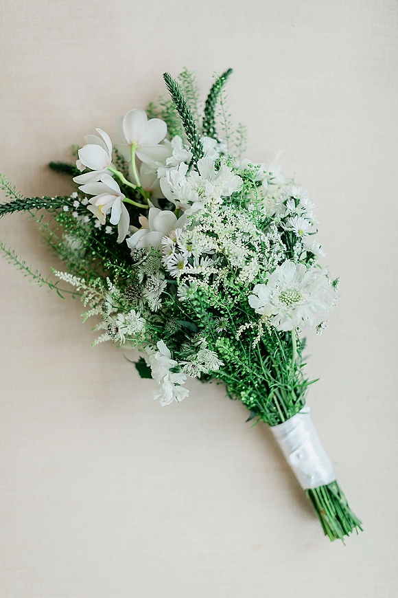 Bridal bouquet with white wildflowers and fern fronds, hand-tied with ribbon wrap and stems against a beige backdrop