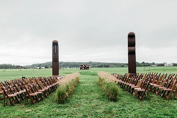 Ceremony setup with outdoor ceremony seating, wood folding chairs and sculptural pillars lining a grass aisle in a rolling field under clouds