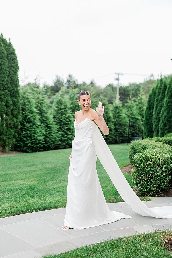 Bridal portrait of a smiling bride in a strapless wedding dress with a long veil on a stone patio beside green hedges and trees